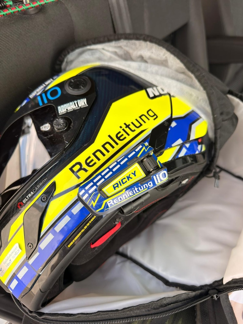 May include: A yellow, blue, and black racing helmet featuring the text Rennleitung, Ricky, and 110. The helmet has a dark visor, carbon fiber detailing, and a prominent yellow side panel with blue accents and white dashed lines.