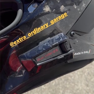 May include: A black motorcycle helmet with a forged carbon fiber pattern, a purple-tinted visor, and a communication device attached to the side. The helmet features the yellow text @extra_ordinary_garage and the red text duc ride 23.