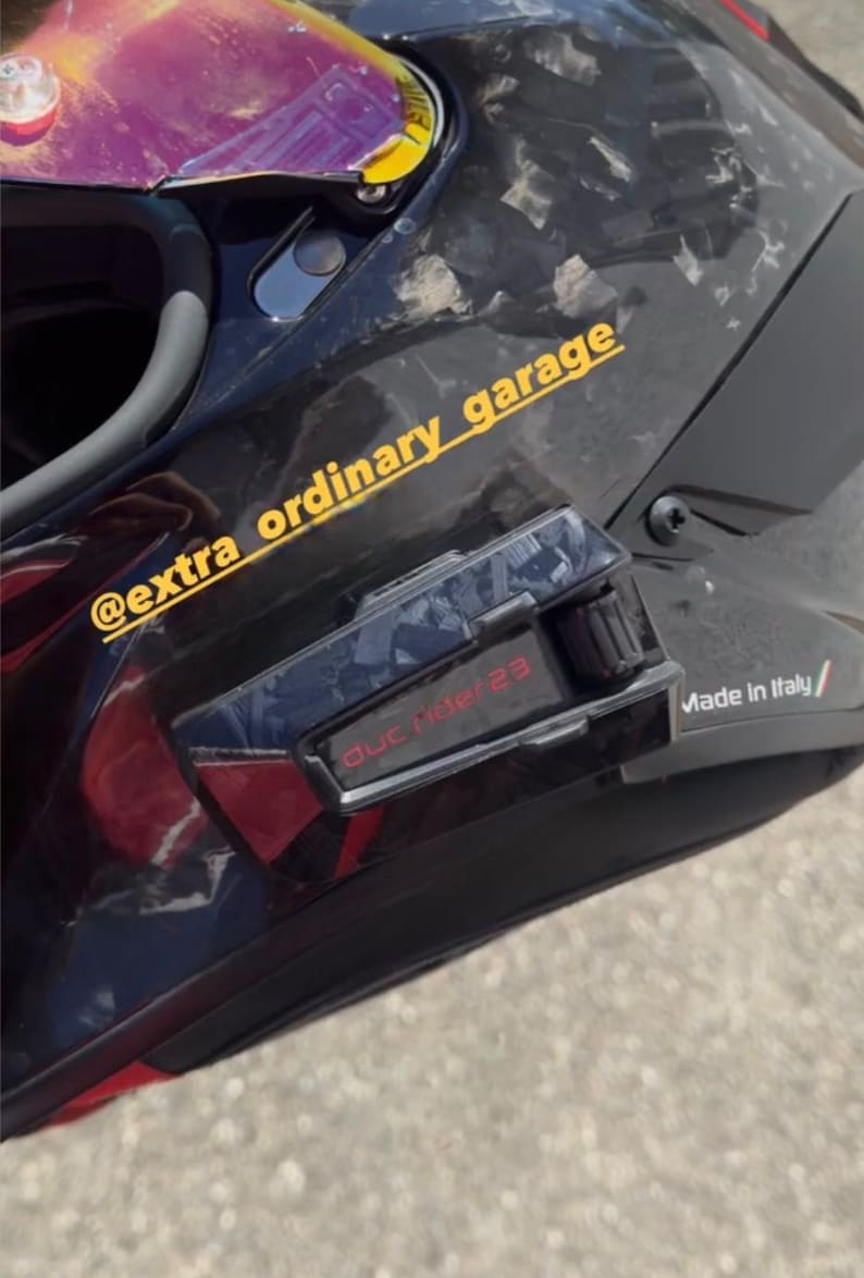 May include: A black motorcycle helmet with a forged carbon fiber pattern, a purple-tinted visor, and a communication device attached to the side. The helmet features the yellow text @extra_ordinary_garage and the red text duc ride 23.