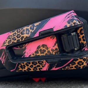 May include: A motorcycle helmet communication device featuring a black plastic housing with a decorative skin. The surface displays a vibrant pink and black brushstroke pattern layered over a brown and black leopard print design with a black dial.