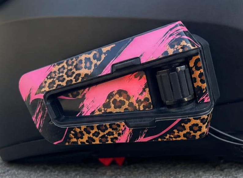 May include: A motorcycle helmet communication device featuring a black plastic housing with a decorative skin. The surface displays a vibrant pink and black brushstroke pattern layered over a brown and black leopard print design with a black dial.