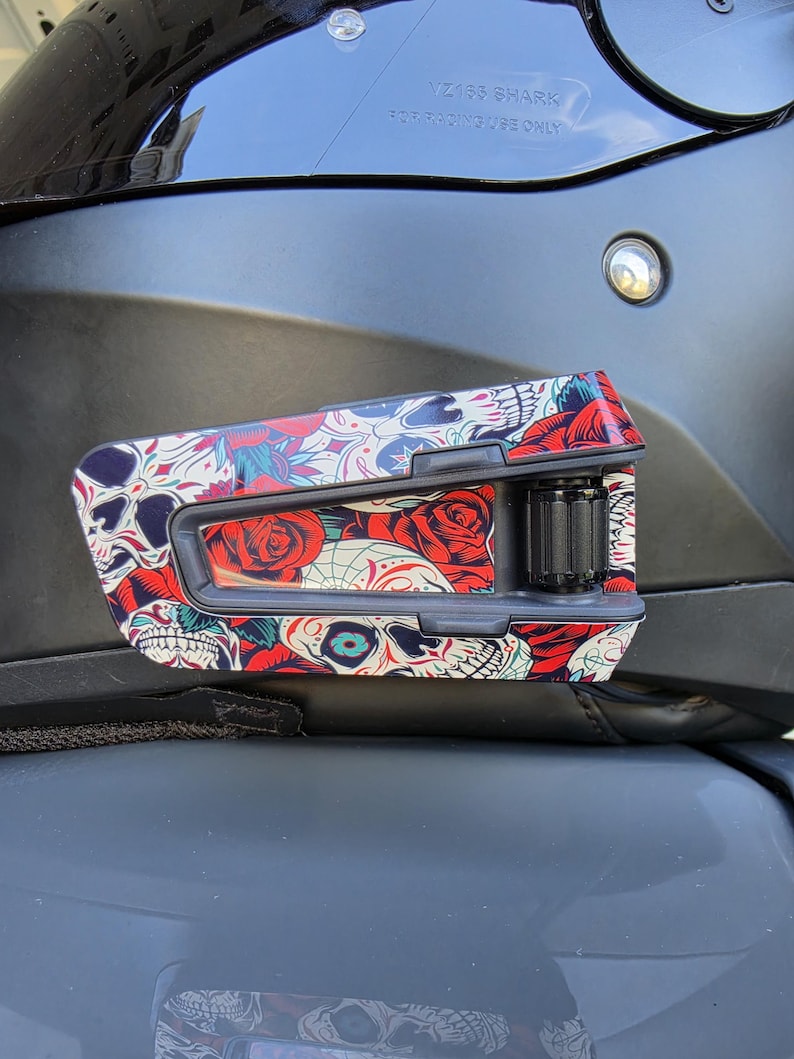 May include: A black motorcycle helmet communication device featuring a vibrant graphic overlay of skulls and red roses. The device is mounted on the side of a matte black helmet, which displays the text VZ160 SHARK FOR RACING USE ONLY.