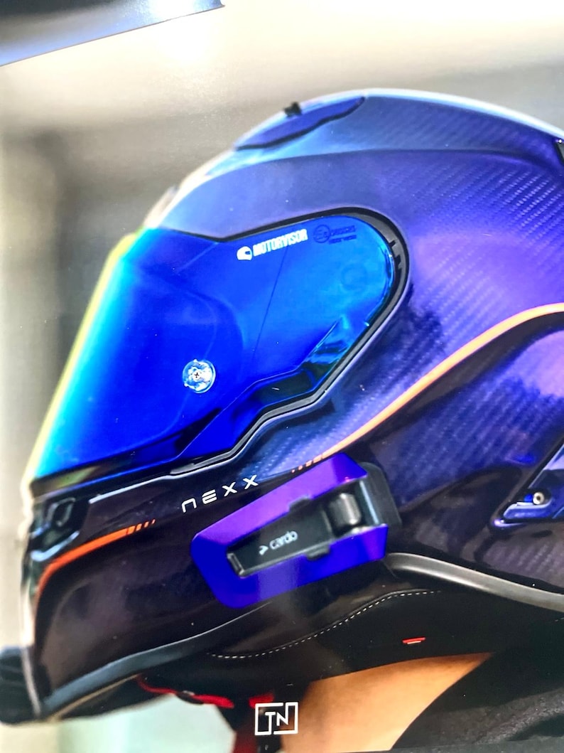 May include: A Nexx motorcycle helmet featuring a deep blue and purple carbon fiber shell with orange accents. The helmet includes a reflective blue visor with Motorvisor branding and a Cardo communication device attached to the side.