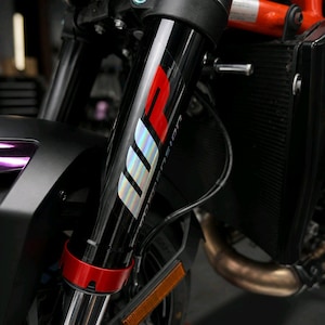 May include: Close-up of a motorcycle front suspension fork. The black fork has a red and silver "WP Suspension" logo. A red ring accents the bottom of the fork. The motorcycle's orange frame and radiator are visible in the background.