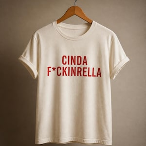 May include: A cream-colored short-sleeve t-shirt with rolled sleeves, featuring the red text "CINDA F*CKINRELLA" printed across the chest. The garment is displayed on a wooden hanger against a neutral background, showcasing a crew neckline.