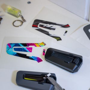 May include: Two black communication headset units for helmets, one with a utility knife blade inserted, alongside adhesive decals featuring a stylized letter A in black, white, red, and yellow, and a separate decal with a vibrant blue and pink pattern.