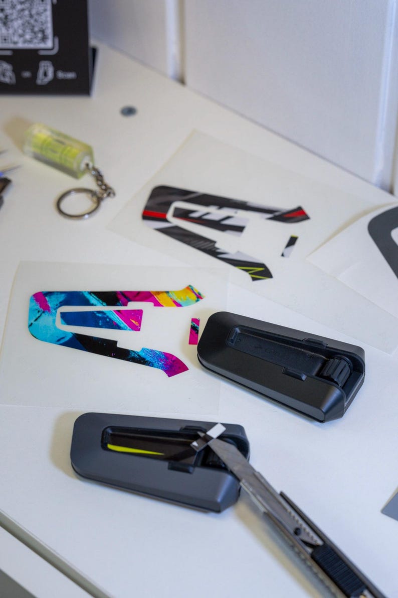 May include: Two black communication headset units for helmets, one with a utility knife blade inserted, alongside adhesive decals featuring a stylized letter A in black, white, red, and yellow, and a separate decal with a vibrant blue and pink pattern.