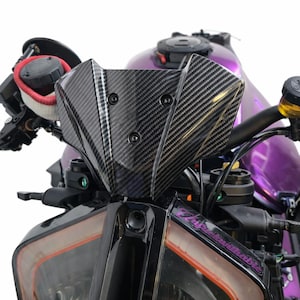 May include: Close-up of a motorcycle's front end, featuring a carbon fiber windscreen and black components. The motorcycle has a purple fuel tank and handlebars. The headlight has an orange outline. The text "Thug Life Unbreakable" is visible on the side.