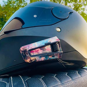 May include: A matte black motorcycle helmet featuring a dark tinted visor and a communication device attached to the side. The device is decorated with a colorful graphic print depicting a Japanese landscape with cherry blossoms, a bridge, and a pagoda.