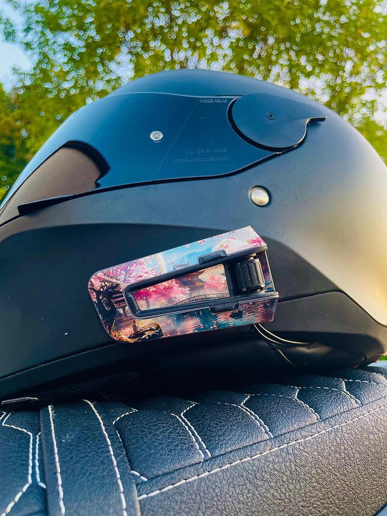 May include: A matte black motorcycle helmet featuring a dark tinted visor and a communication device attached to the side. The device is decorated with a colorful graphic print depicting a Japanese landscape with cherry blossoms, a bridge, and a pagoda.
