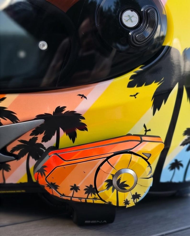 Könnte beinhalten: Ein Motorradhelm mit einem lebendigen tropischen Sonnenuntergangsmotiv, schwarzen Palmensilhouetten, orangefarbenen und gelben Diagonalstreifen sowie kleinen Vögeln. Ein passendes Kommunikationsgerät ist an der Seite angebracht und zeigt dasselbe Muster.
