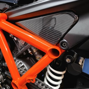May include: A motorcycle frame featuring a bright orange tubular steel structure, a triangular carbon fibre side panel, a white coil-over shock absorber, and a metallic drive chain. The mechanical components are visible behind the frame.
