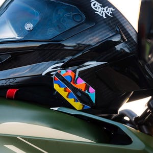 May include: A black carbon fiber motorcycle helmet with a glossy finish and a white logo, featuring a small, colorful, abstract-patterned geometric accessory attached to the side, resting on a matte olive green motorcycle fuel tank with white text.