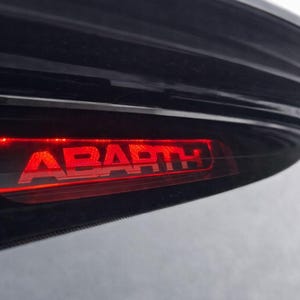 Peut inclure: Un troisième feu stop automobile noir présentant le mot ABARTH illuminé par une lumière LED rouge vif. Le boîtier est sombre et élégant, intégré dans la carrosserie incurvée d'un véhicule, mettant en valeur le texte rouge lumineux.