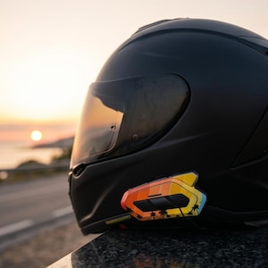 May include: A matte black full-face motorcycle helmet featuring a tinted visor and a mounted communication device on the side. The device displays a vibrant gradient pattern transitioning from yellow to orange and blue, accented with black palm tree silhouettes.