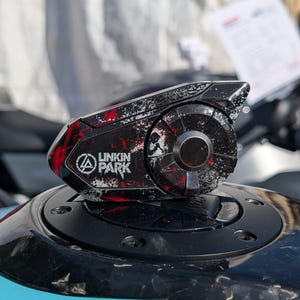May include: A black motorcycle helmet communication device featuring a Linkin Park logo and a distressed graphic pattern in red, white, and black. The unit has a circular control dial on the side and a sleek, angular housing design.