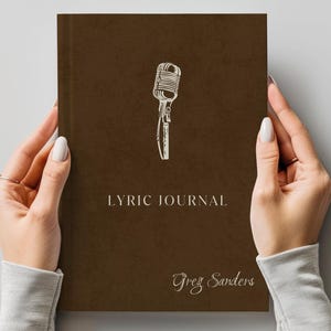 May include: A brown lyric journal with a microphone illustration and the text "Lyric Journal" and "Greg Sanders" in a handwritten style font. The journal is held by two hands.