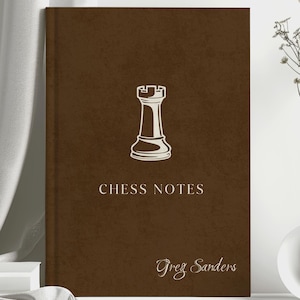 May include: A brown notebook featuring a rook chess piece graphic and the words "CHESS NOTES". The name "Greg Sanders" is printed at the bottom. A vase with dried flowers is to the right.