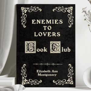 May include: A black book cover with the title "Enemies to Lovers Book Club" in white script. The cover has decorative flourishes and the author's name, Elizabeth Ann Montgomery, at the bottom. A clear vase with dried flowers sits to the right.
