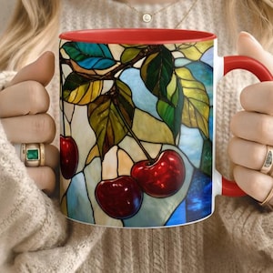 Peut inclure: Mug en céramique blanche avec intérieur et anse rouges. Le mug présente un motif de vitrail de cerises sur une branche avec des feuilles vertes et un fond bleu et jaune. Les cerises sont d'un rouge profond.