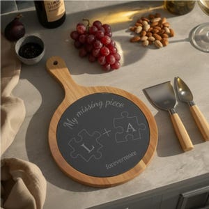 Puede incluir: Una tabla de quesos de madera con un centro de pizarra gris oscuro, con el texto "My missing piece" y piezas de rompecabezas con las letras "L" y "A". Incluye cuchillos para queso y uvas.