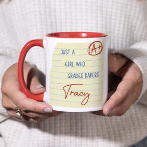 May include: A white ceramic mug with a red interior and handle. The mug features text that reads "Just a girl who grades papers" with the name "Tracy" and an A+ grade mark.