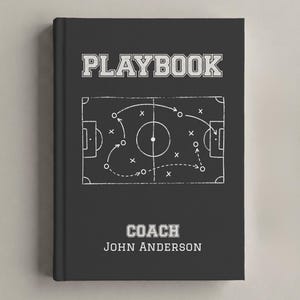 May include: A black hardback book titled PLAYBOOK with a white graphic of a football pitch diagram featuring arrows, circles, and x marks. Below the diagram, the text reads COACH and JOHN ANDERSON in white block lettering on the front cover.