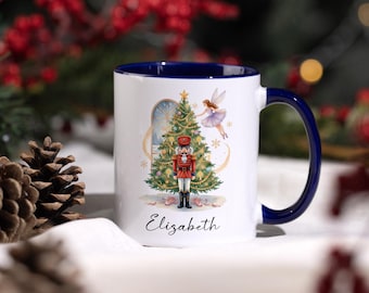 May include: White ceramic mug with a blue interior and handle. The mug features a Christmas tree, a nutcracker, and a fairy illustration. The name "Elizabeth" is written in script below the illustration.