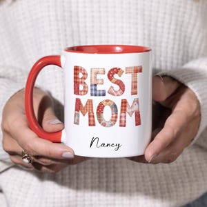 May include: White ceramic mug with a red interior and handle. The mug features the words "BEST MOM" in patchwork-style letters and the name "Nancy" in a script font.