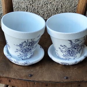 Può includere: Due vasi da fiori in ceramica bianca con sottovasi abbinati, ciascuno caratterizzato da un dettagliato motivo floreale blu. I vasi hanno un bordo largo e un corpo affusolato, con illustrazioni botaniche blu che avvolgono la superficie esterna dei vasi e dei sottovasi.