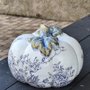 Peut inclure: Une citrouille décorative blanche ornée d'un motif floral bleu de type toile de Jouy. La tige et les feuilles sculptées présentent une finition patinée bleu et or métallique, mettant en valeur les détails texturés des éléments végétaux.