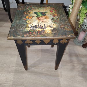 Op de afbeelding: Een vierkante houten bijzettafel met vier zwarte poten en een bovenblad met een gedetailleerde illustratie van een persoon met een groene hoed die schaakt, omringd door klokken, vlinders en bloemen. De rand is versierd met gouden kaartsymbolen.