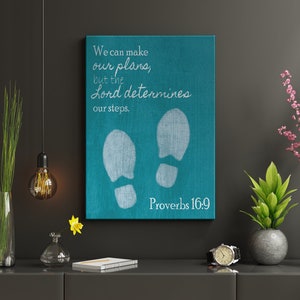 Puede incluir: Lienzo de pared de color turquesa con texto blanco que dice "Podemos hacer nuestros planes, pero el Señor determina nuestros pasos. Proverbios 16:9". Se ven dos huellas blancas en el lienzo.