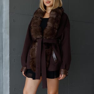 Peut inclure: Un manteau marron foncé avec un col et des poignets en fourrure, et une ceinture assortie. Le manteau est porté sur une tenue noire. Le fond est un mur gris.