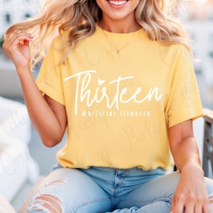 May include: A yellow t-shirt with the text "Thirteen #OFFICIAL TEENAGER" in white. The shirt is being worn by a person with long blonde hair.