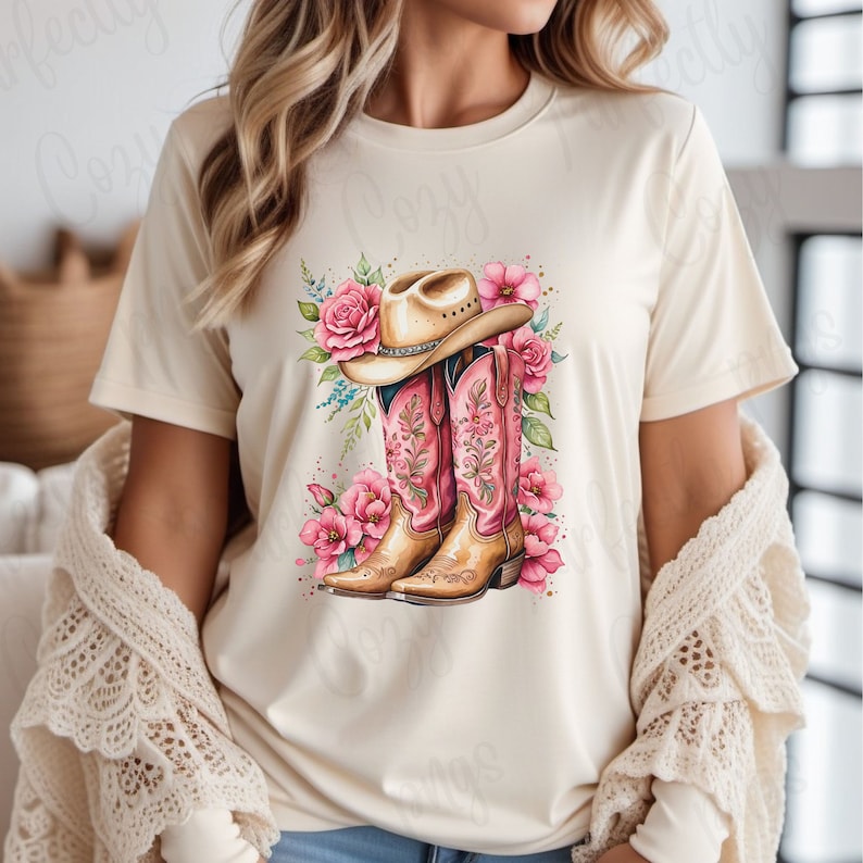 K&ouml;nnte beinhalten: Ein wei&szlig;es T-Shirt mit einem Grafikdesign von rosa Cowboystiefeln und einem braunen Cowboyhut mit Blumenmuster. Die Stiefel haben rosa Stickereien und die Blumen sind rosa und wei&szlig;.
