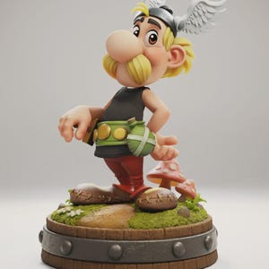 Op de afbeelding: Gedetailleerd beeldje van een stripfiguur met een grote neus, gele snor en gevleugelde helm. Het figuur draagt een zwarte top, rode broek en een groene riem met een buidel. Het staat op een houten voet met paddenstoel- en grasdetails. Figuur van Tanuke.