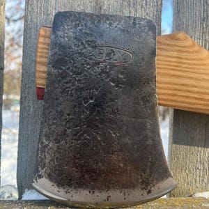 May include: Close-up of a vintage axe head with the text "KELLY" and "WORLDS FINEST" stamped on the metal. The axe head is dark gray with a slightly curved blade. The wooden handle is visible in the background.