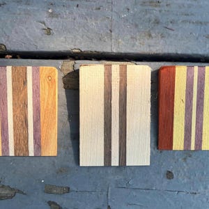 May include: Three rectangular wooden blocks featuring vertical stripes of varying wood tones. The left block has light, medium, and purple-toned wood. The center block has light and dark brown stripes. The right block has red, yellow, and purple stripes.