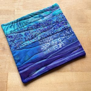 May include: A square, quilted fabric item with a gradient of blue hues. The design features horizontal bands of varying patterns and shades, from teal to deep indigo, stitched together. The edges are neatly finished.