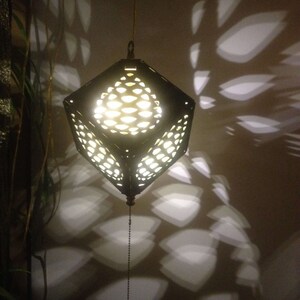 Op de afbeelding: Een zwarte hanglamp van metaal met een geometrisch ontwerp werpt ingewikkelde schaduwen op de muur. De lamp is aan en straalt een warm licht uit.