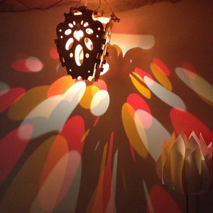 Op de afbeelding: Een houten lampenkap met ingewikkelde uitsnijdingen werpt kleurrijke, abstracte schaduwen op een muur. De schaduwen zijn in tinten rood, oranje, geel en wit.