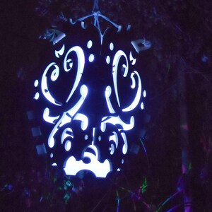 Op de afbeelding: Een zwarte metalen lantaarn met een wit licht dat schijnt door ingewikkelde uitgesneden ontwerpen. De lantaarn hangt aan een tak.