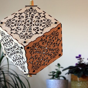 Op de afbeelding: Een houten kubus lampenkap met ingewikkelde lasergesneden ontwerpen. De lampenkap is gemaakt van licht hout en heeft een bruine vlek. De lampenkap hangt aan het plafond.