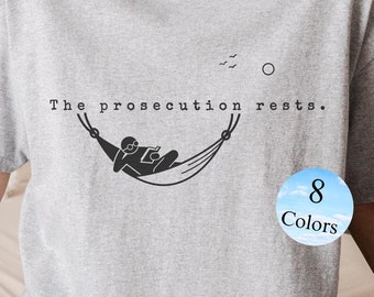 以下が含まれることがあります： 「The prosecution rests.」というテキストと、ハンモックでリラックスしている人のグラフィックがプリントされたグレーのTシャツ。グラフィックは白黒です。このTシャツは8色展開です。