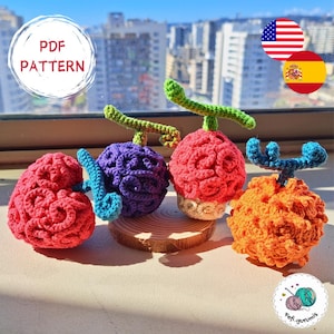 Może przedstawiać: Cztery szydełkowe rzeźby owoców w kolorach czerwonym, fioletowym i pomarańczowym, każda z zieloną lub niebieską łodygą, są wystawione. Obraz zawiera tekst "PDF PATTERN" i logo z przędzą i igłami. Owoce są wystawione na drewnianej podstawie.