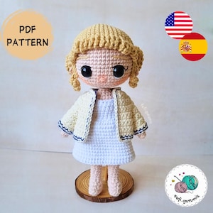Puede incluir: Muñeca de ganchillo hecha a mano con chaqueta amarilla clara, vestido blanco y pelo rubio. La muñeca tiene grandes ojos negros y está sobre una base de madera. La imagen incluye el texto "PDF PATTERN".