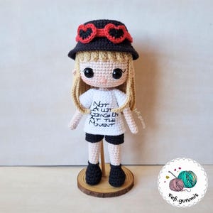 Op de afbeelding: Een handgemaakte gehaakte pop met blond haar, een zwarte hoed met rode hartvormige accenten, een wit t-shirt met zwarte tekst en een zwarte korte broek. De pop staat op een houten voet.
