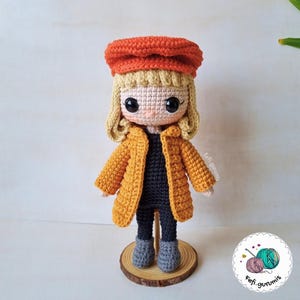 Op de afbeelding: Een handgemaakte gehaakte pop met blond haar, een oranje baret en een mosterdgele jas. De pop heeft een zwarte top, een grijze broek en grijze laarzen. Hij staat op een houten voet.