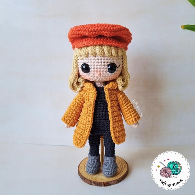 Op de afbeelding: Een handgemaakte gehaakte pop met blond haar, een oranje baret en een mosterdgele jas. De pop heeft een zwarte top, een grijze broek en grijze laarzen. Hij staat op een houten voet.